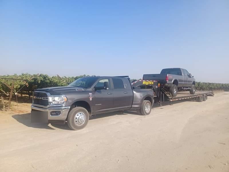 A pickup hauling another truck on a gooseneck trailer beside a vineyard